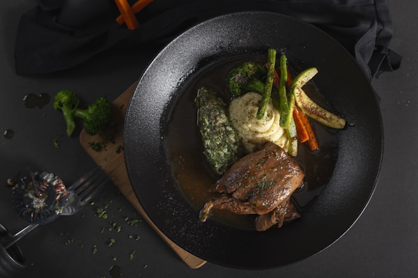 A dark ceramic plate features an elegantly arranged meal consisting of a piece of braised meat, mashed potatoes, grilled zucchini, carrots, and broccoli. The dish is garnished with herbs and surrounded by a rich sauce. A rustic wooden board serves as a base, against a dark background with scattered green herbs and a partially visible fork.