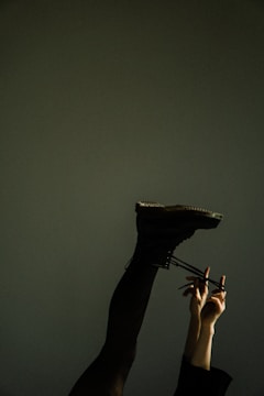 A person's leg is extended upwards against a muted background. The leg is clad in dark pants and a black boot. A pair of hands is seen tightening the laces of the boot.