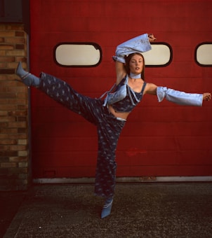 A dancer poses in an expressive stance against a vibrant red garage door. She is wearing a distinctive blue denim outfit with flared sleeves and matching pants, adding a modern twist to her look. The brick wall on one side complements the industrial setting.