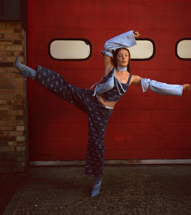 A dancer poses in an expressive stance against a vibrant red garage door. She is wearing a distinctive blue denim outfit with flared sleeves and matching pants, adding a modern twist to her look. The brick wall on one side complements the industrial setting.