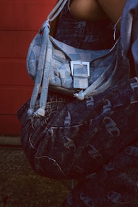 A close-up view of a person wearing denim clothing, featuring a light blue shoulder bag with a prominent silver buckle. The background consists of a red wall, adding contrast to the image.