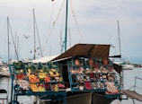 A floating market stall displays a variety of colorful fruits and vegetables in blue crates. There are other items such as pottery, herbs, and various packaged products. The stall is set aboard a boat docked at a marina with multiple sailboats in the background, and a gray sky overhead. Several cloth canopies provide shade for the stall.