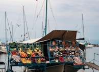 A floating market stall displays a variety of colorful fruits and vegetables in blue crates. There are other items such as pottery, herbs, and various packaged products. The stall is set aboard a boat docked at a marina with multiple sailboats in the background, and a gray sky overhead. Several cloth canopies provide shade for the stall.