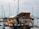 A floating market stall displays a variety of colorful fruits and vegetables in blue crates. There are other items such as pottery, herbs, and various packaged products. The stall is set aboard a boat docked at a marina with multiple sailboats in the background, and a gray sky overhead. Several cloth canopies provide shade for the stall.