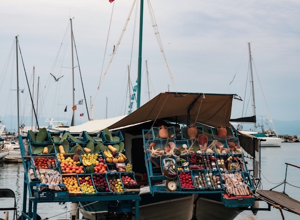 A floating market stall displays a variety of colorful fruits and vegetables in blue crates. There are other items such as pottery, herbs, and various packaged products. The stall is set aboard a boat docked at a marina with multiple sailboats in the background, and a gray sky overhead. Several cloth canopies provide shade for the stall.