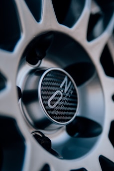 Close-up view of a car wheel rim with a focus on the center cap, which has a carbon fiber pattern and a logo imprinted on it. The design is intricate, with metallic spokes radiating from the center.