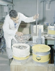 A person wearing a white lab coat and glasses is using a scoop to pour a large number of metal coins into a yellow container lined with a white cloth. The setting appears to be an industrial or laboratory environment, with metal equipment and other containers in the background.
