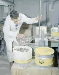 A person wearing a white lab coat and glasses is using a scoop to pour a large number of metal coins into a yellow container lined with a white cloth. The setting appears to be an industrial or laboratory environment, with metal equipment and other containers in the background.