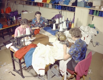 Several people are sitting at sewing machines, busy working with various fabrics. The workspace is filled with colorful threads and cloth materials, indicating a garment workshop or sewing room.