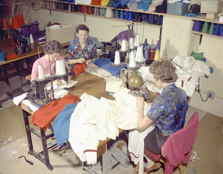 Several people are sitting at sewing machines, busy working with various fabrics. The workspace is filled with colorful threads and cloth materials, indicating a garment workshop or sewing room.