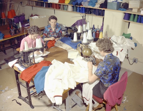 Several people are sitting at sewing machines, busy working with various fabrics. The workspace is filled with colorful threads and cloth materials, indicating a garment workshop or sewing room.
