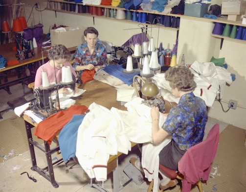 Several people are sitting at sewing machines, busy working with various fabrics. The workspace is filled with colorful threads and cloth materials, indicating a garment workshop or sewing room.