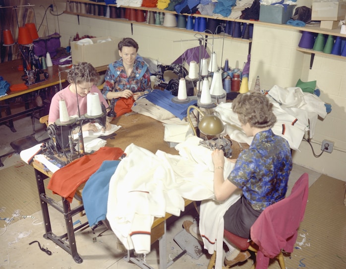Several people are sitting at sewing machines, busy working with various fabrics. The workspace is filled with colorful threads and cloth materials, indicating a garment workshop or sewing room.