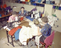 Several people are sitting at sewing machines, busy working with various fabrics. The workspace is filled with colorful threads and cloth materials, indicating a garment workshop or sewing room.