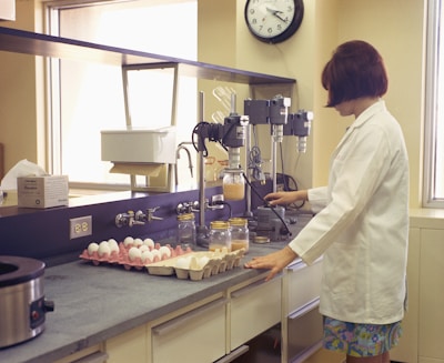 A laboratory setting with a person wearing a white lab coat working at a counter. There are trays of eggs, jars, and laboratory equipment, including a mixer and a clock on the wall.