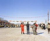 Several people stand near a start and finish line for a snowmobile race on a snow-covered track. Spectators gather around, some seated on a platform to the left. A line of yellow snowmobiles waits for the race to begin. Bright blue sky overhead with orange and blue pennant flags strung across the track.