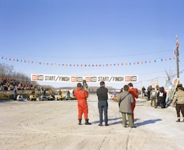 Several people stand near a start and finish line for a snowmobile race on a snow-covered track. Spectators gather around, some seated on a platform to the left. A line of yellow snowmobiles waits for the race to begin. Bright blue sky overhead with orange and blue pennant flags strung across the track.