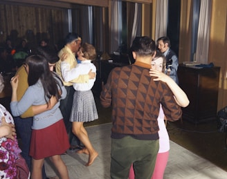Several couples are dancing closely in a warmly lit room with wooden paneling and large windows. In the background, a musician is playing a keyboard, adding to the intimate atmosphere. The dancers appear relaxed and engaged in conversation.