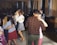 Several couples are dancing closely in a warmly lit room with wooden paneling and large windows. In the background, a musician is playing a keyboard, adding to the intimate atmosphere. The dancers appear relaxed and engaged in conversation.