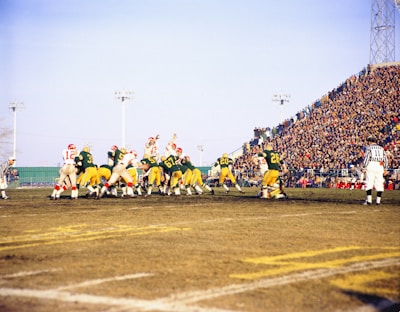 A football game is being played on a field with teams wearing green and yellow, and red and white uniforms. Players are actively engaged, some in mid-action, under a clear blue sky. A large crowd of spectators fills the stands on the right side, and referees are visible on the sidelines.