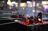 A close-up view of skewered, grilled meat with a charred, smoky appearance. The meat is positioned over a grill with glowing red coals visible beneath, suggesting a cooking process in progress. The background is blurred, filled with bokeh lights, indicating a nighttime or indoor setting.