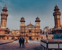 An ornate historical mosque featuring intricate architectural details, including minarets and decorative tile work. The building is framed by a clear blue sky, and there are several people visible in the courtyard, including two in the foreground. The structure is undergoing some restoration or maintenance work, as evidenced by the scaffolding present.