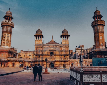 An ornate historical mosque featuring intricate architectural details, including minarets and decorative tile work. The building is framed by a clear blue sky, and there are several people visible in the courtyard, including two in the foreground. The structure is undergoing some restoration or maintenance work, as evidenced by the scaffolding present.