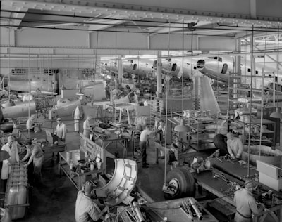A busy aircraft manufacturing facility with numerous workers engaged in assembling and constructing large components of airplanes. The factory is equipped with various workbenches, tools, and materials scattered throughout the spacious interior. Workers are wearing uniforms and appear to be focused on their tasks around partially assembled aircraft sections.