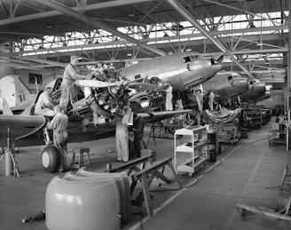 Several workers are engaged in the maintenance and assembly of aircraft inside an expansive hangar. They are working on multiple planes, focusing on parts such as engines and propellers. The hangar is spacious with large windows and overhead steel beams, and there are various tools, workbenches, and equipment scattered throughout the workspace.
