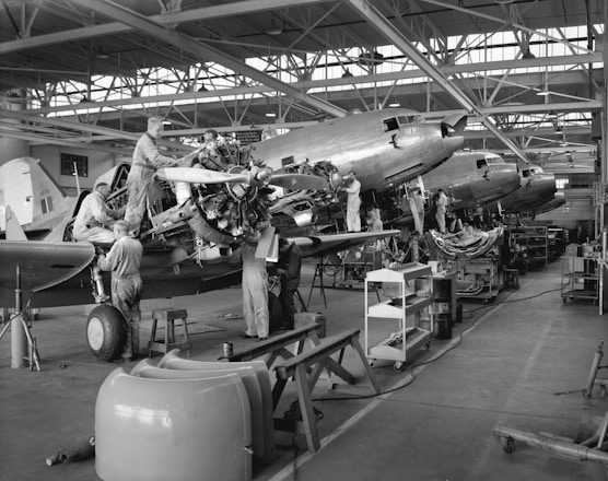 Several workers are engaged in the maintenance and assembly of aircraft inside an expansive hangar. They are working on multiple planes, focusing on parts such as engines and propellers. The hangar is spacious with large windows and overhead steel beams, and there are various tools, workbenches, and equipment scattered throughout the workspace.