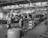 Several workers are engaged in the maintenance and assembly of aircraft inside an expansive hangar. They are working on multiple planes, focusing on parts such as engines and propellers. The hangar is spacious with large windows and overhead steel beams, and there are various tools, workbenches, and equipment scattered throughout the workspace.