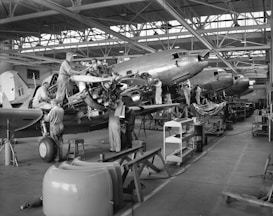 Several workers are engaged in the maintenance and assembly of aircraft inside an expansive hangar. They are working on multiple planes, focusing on parts such as engines and propellers. The hangar is spacious with large windows and overhead steel beams, and there are various tools, workbenches, and equipment scattered throughout the workspace.