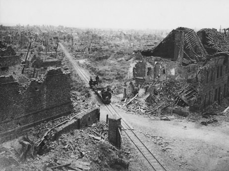 A vast landscape of destruction marked by ruined buildings, shattered roofs, and debris scattered throughout the area. The remnants of a railway track extend through the destruction, with a few figures silhouetted against the devastation. The scene reflects the aftermath of a significant conflict, with no signs of life or vegetation.