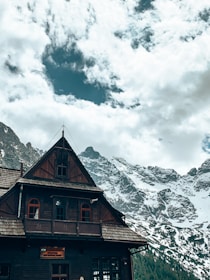 A wooden chalet-style building with a steep roof and several small windows is set against a backdrop of snow-capped mountains. The sky above is partly cloudy, with patches of blue visible through the clouds.