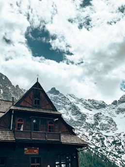 A wooden chalet-style building with a steep roof and several small windows is set against a backdrop of snow-capped mountains. The sky above is partly cloudy, with patches of blue visible through the clouds.