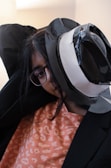 A person wearing glasses appears to be dressed in a black suit with a white and black hat. The head is tilted at an unusual angle, creating a playful and surreal look. The person is also wearing an orange patterned shirt.