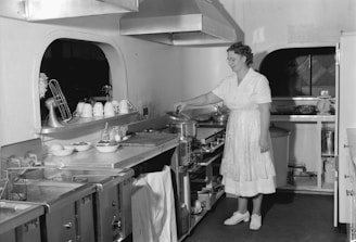 A woman in a vintage kitchen is cooking at a stove, wearing a white uniform and apron. The kitchen is equipped with various utensils, dishes, and appliances. The countertop is organized with cups hanging next to a mixer and bowls filled with ingredients. The walls have a rounded design, and there is a large industrial exhaust fan above the cooking area.