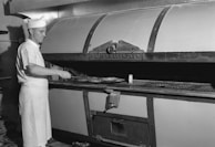 A baker dressed in white uniform and cap uses a long-handled peel to place or retrieve bread from a large industrial oven labeled 'Master Baker Oven'. The setting appears to be a bakery or a baking manufacturing facility.