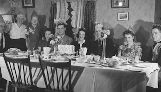 A black-and-white photograph showing a group of people seated and standing around a dining table. They are wearing paper party hats and appear to be celebrating a special occasion. The table is set with a variety of dishes, including a large cake in the center. The setting is an indoor dining room with vintage wallpaper and framed art on the walls.