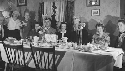A black-and-white photograph showing a group of people seated and standing around a dining table. They are wearing paper party hats and appear to be celebrating a special occasion. The table is set with a variety of dishes, including a large cake in the center. The setting is an indoor dining room with vintage wallpaper and framed art on the walls.