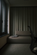 A dimly lit room features a simple, modern sofa with a patterned cushion on it. To the side, a window allows limited light to enter, casting soft shadows. A curtain runs along the back wall, and part of an office chair is visible in the foreground.