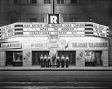 A black and white photograph of a vintage theater with a neon marquee advertising a movie titled 'The More the Merrier' starring Jean Arthur, Joel McCrea, and Charles Coburn. Seven people in matching attire are standing in a line in front of the theater entrance.