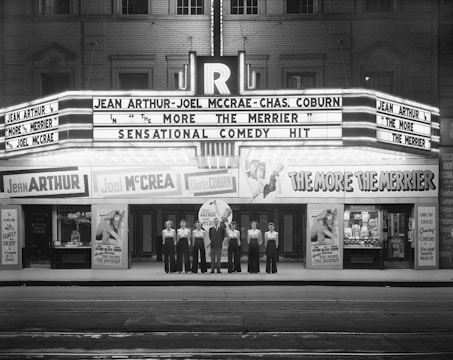 A black and white photograph of a vintage theater with a neon marquee advertising a movie titled 'The More the Merrier' starring Jean Arthur, Joel McCrea, and Charles Coburn. Seven people in matching attire are standing in a line in front of the theater entrance.