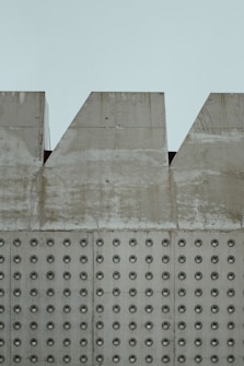 A section of a concrete wall with a pattern of evenly spaced circular indentations. The upper part of the wall has triangular cutouts along its edge. The surface of the wall appears weathered, with visible stains and discoloration.