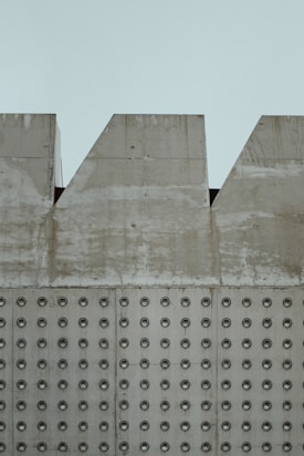 A section of a concrete wall with a pattern of evenly spaced circular indentations. The upper part of the wall has triangular cutouts along its edge. The surface of the wall appears weathered, with visible stains and discoloration.
