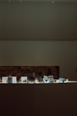 A dimly lit museum exhibit with various small artifacts displayed on a flat surface. A large, abstract artwork is partially visible on the wall behind the exhibit.