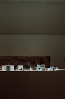 A dimly lit museum exhibit with various small artifacts displayed on a flat surface. A large, abstract artwork is partially visible on the wall behind the exhibit.
