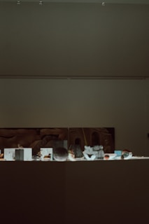 A dimly lit museum exhibit with various small artifacts displayed on a flat surface. A large, abstract artwork is partially visible on the wall behind the exhibit.