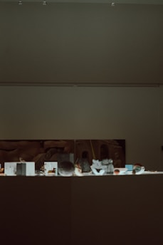A dimly lit museum exhibit with various small artifacts displayed on a flat surface. A large, abstract artwork is partially visible on the wall behind the exhibit.