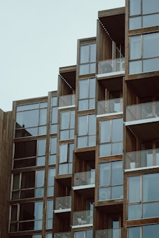 A modern building facade with multiple glass balconies arranged in a stepped pattern. The structure features a combination of wood and glass elements, creating a contemporary design.